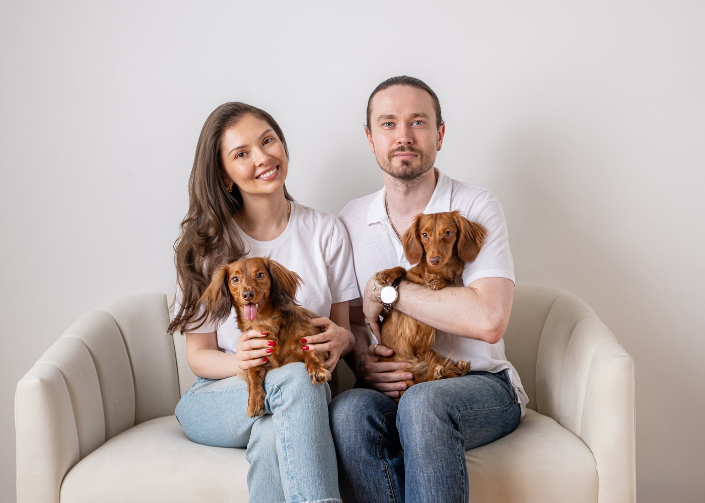 The founders of Nicole & Nora sitting on a cream sofa holding their two long-haired mini dachshunds — Nicole on the left and Nora on the right